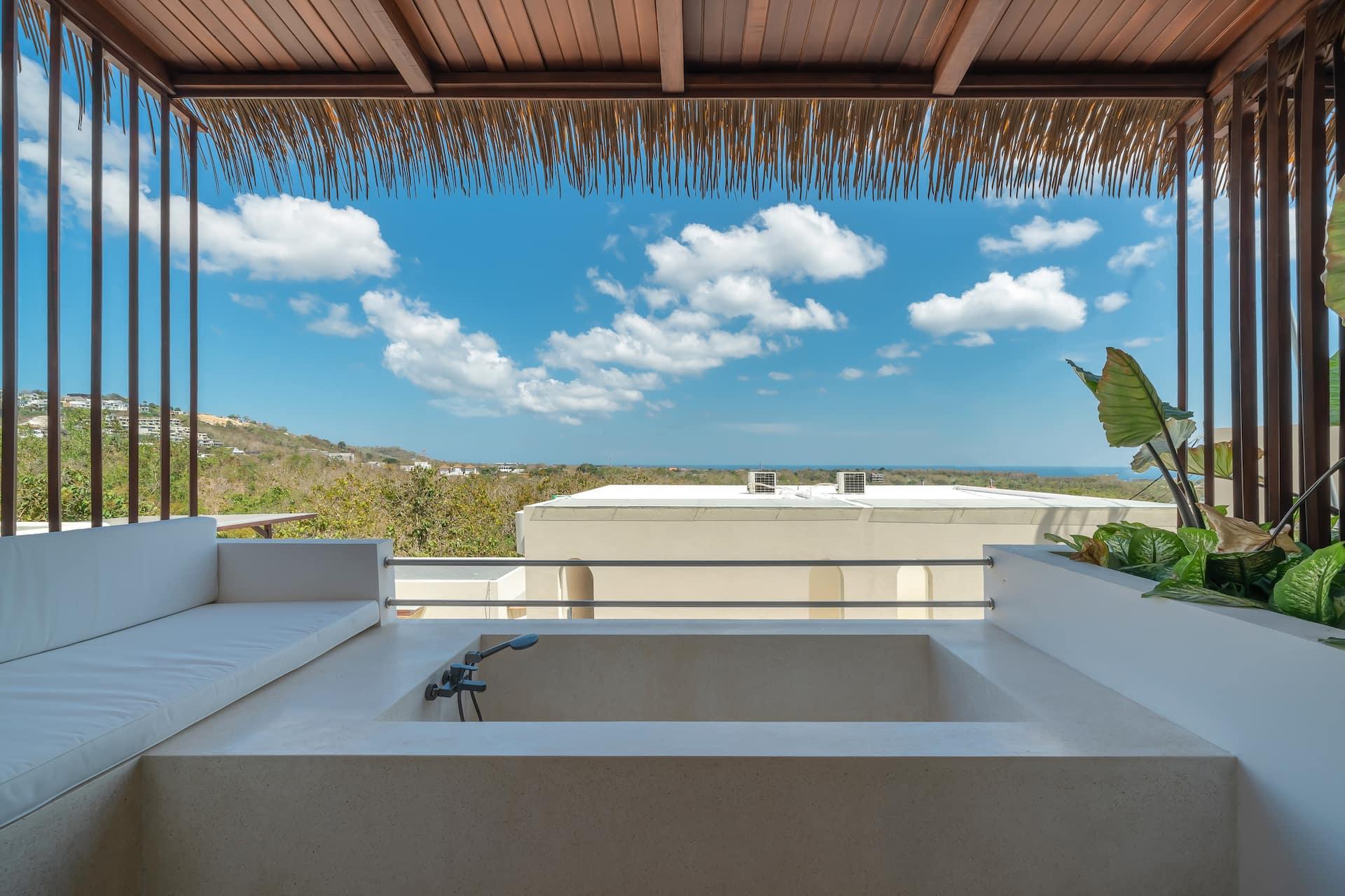 Balcony bathtub and cushioned bench under a thatched roof, looking over green hills to the ocean