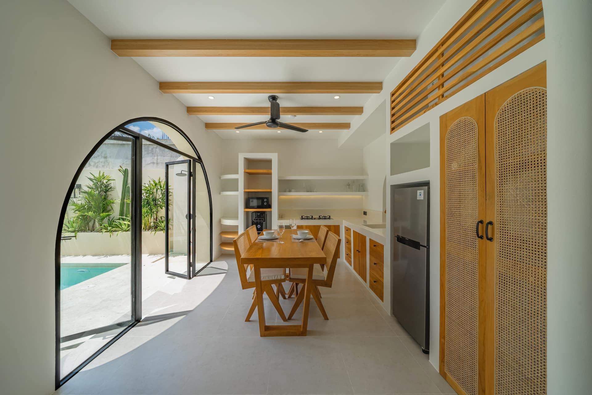 Dining table with rattan chairs beside tall arched glass doors open to the pool