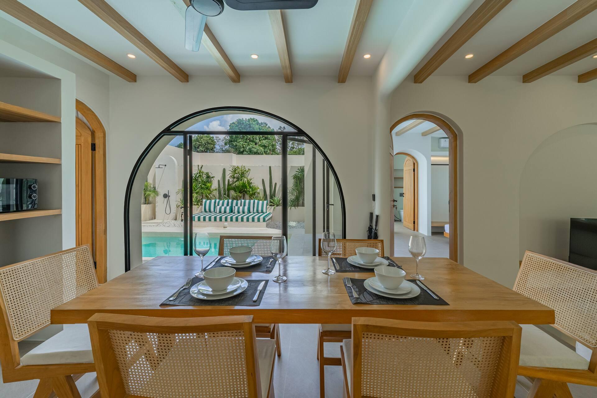 Timber dining table for six with cane chairs, looking out through an arched door to the pool