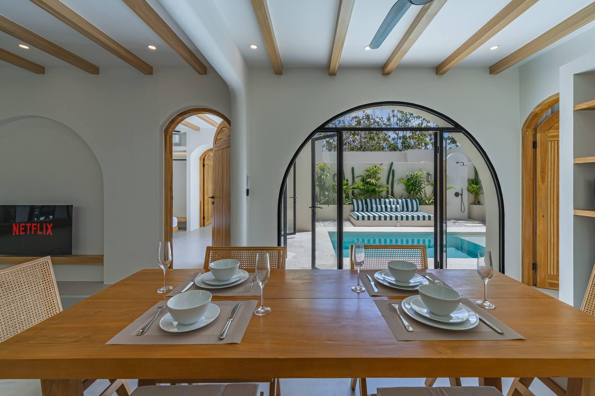 Dining table set for dinner, looking through a black-framed arched door to the pool and daybed