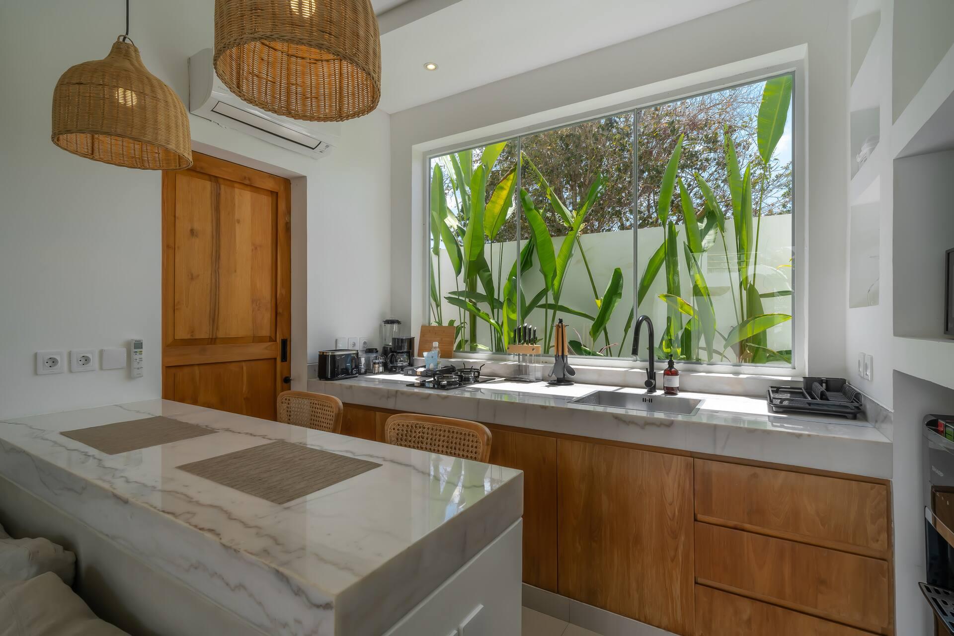 Stone-topped kitchen island in pale timber at Las Casas 5, lit by a window onto the planting