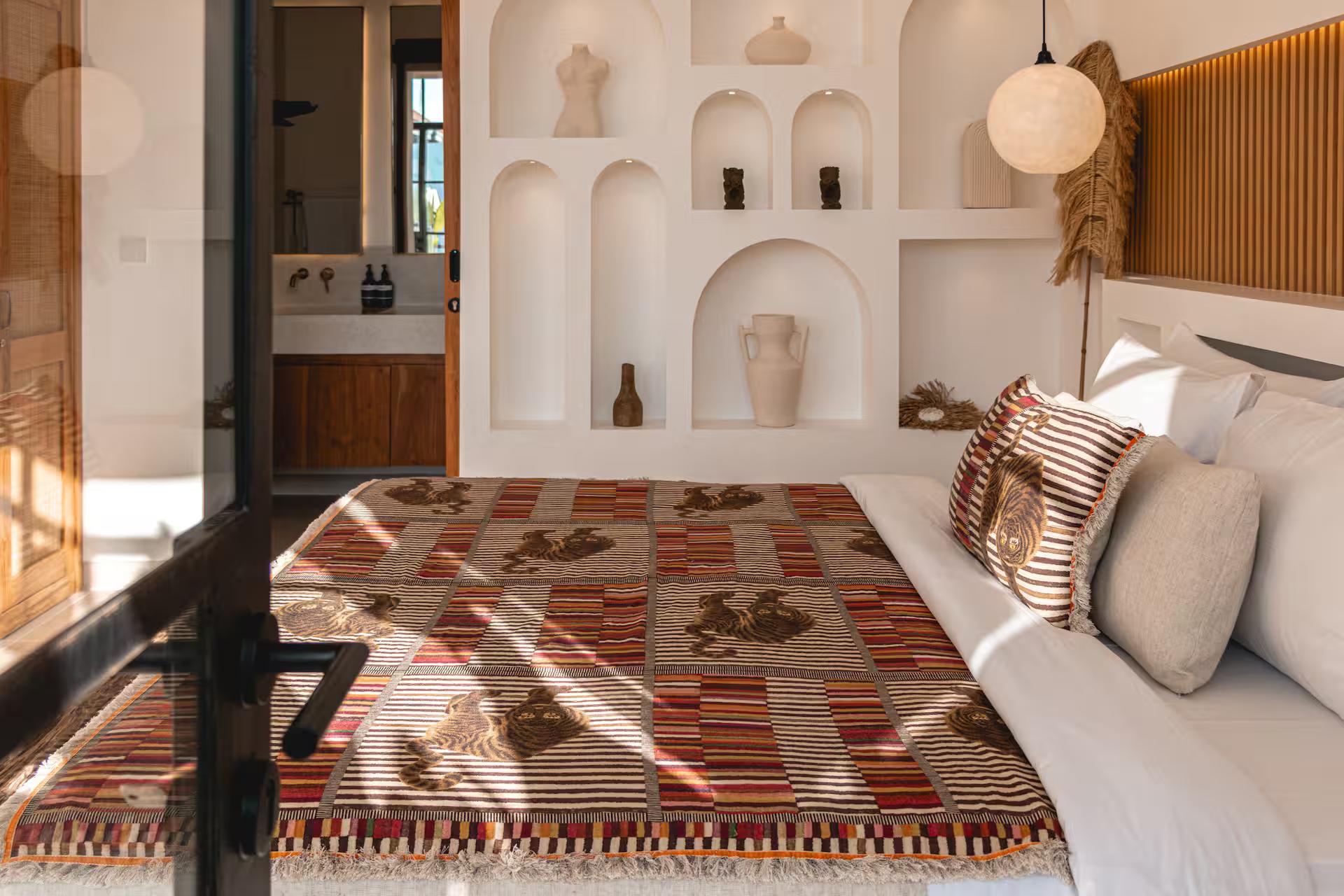 Bed with a kilim throw below arched niches of ceramics, the bathroom visible through the doorway