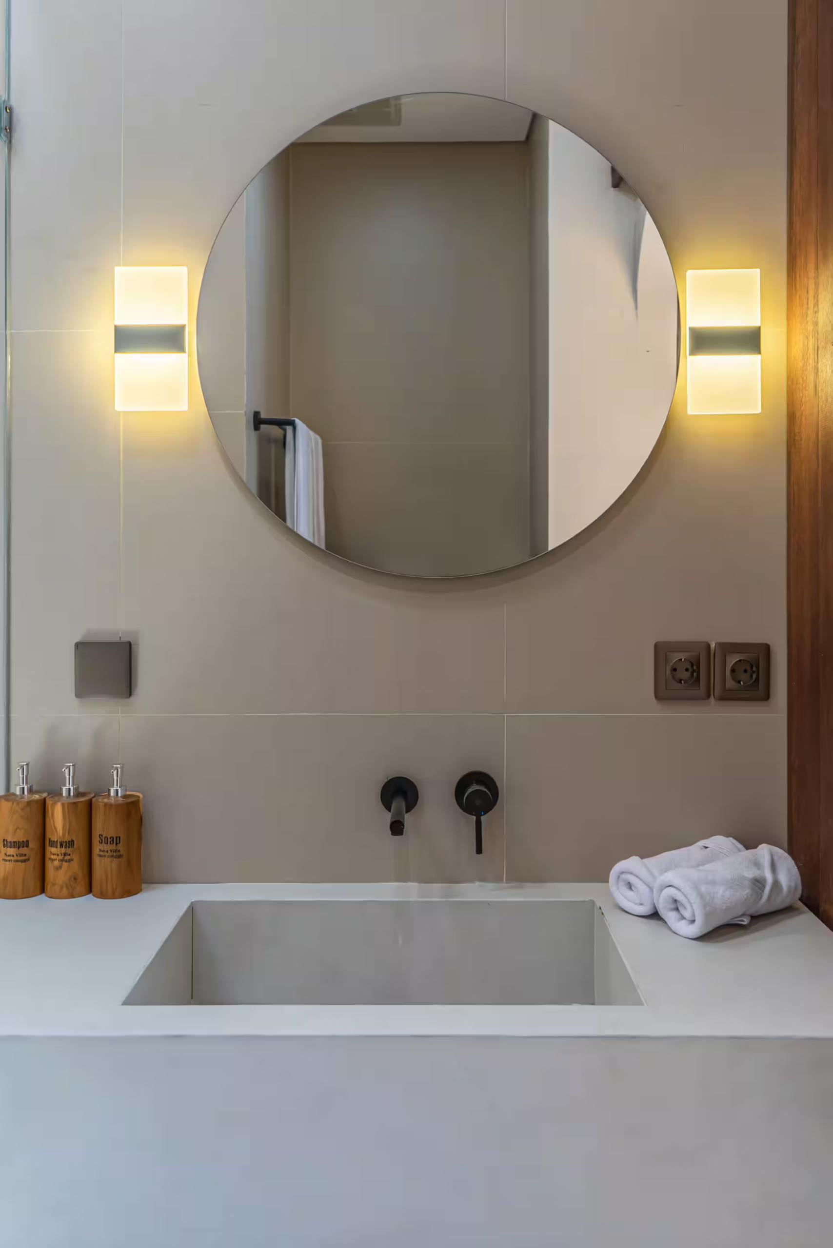 Bathroom with a round mirror between vertical sconces above a concrete trough basin and sunken bath